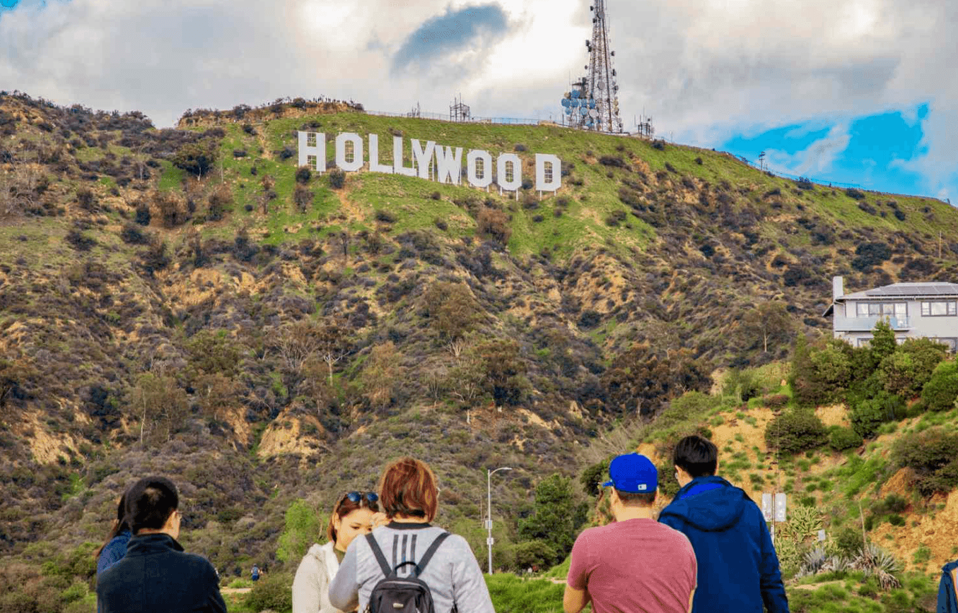 People viewing the iconic Hollywood sign on a green hillside under a partly cloudy sky.